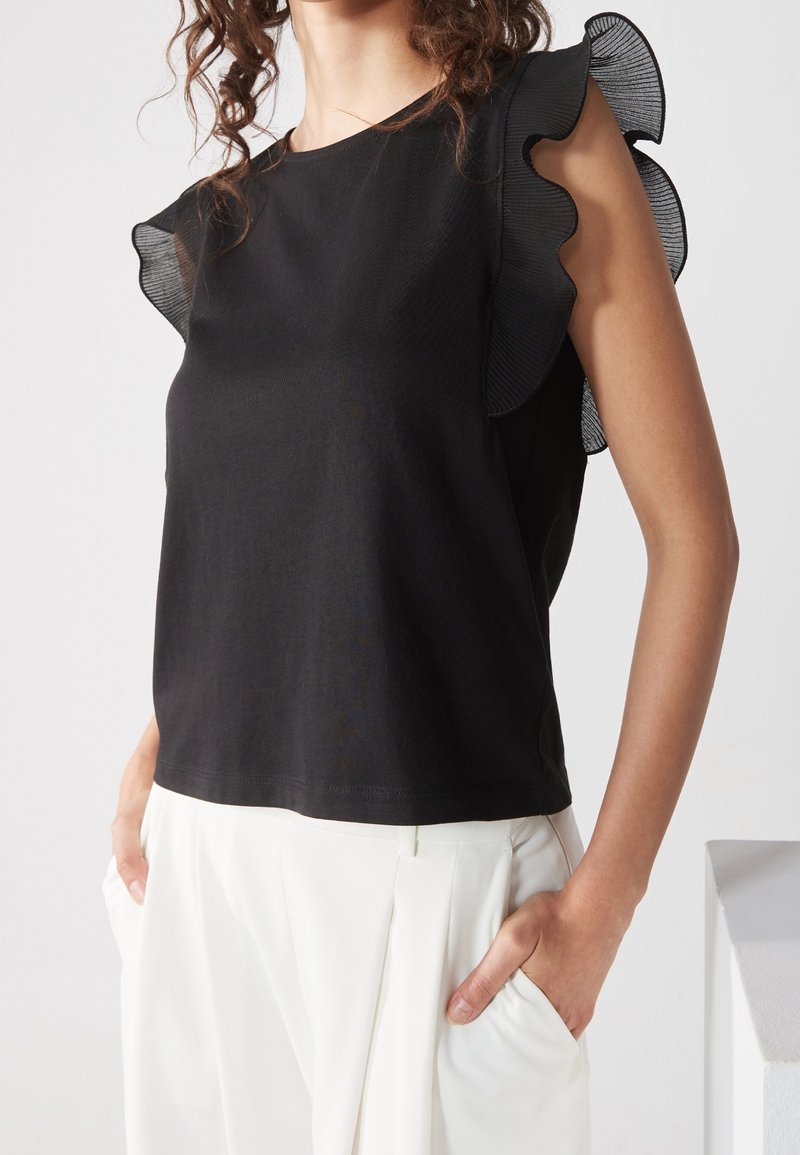 Femme portant un top noir sans manches avec un tissu transparent froncé aux épaules et un pantalon blanc plissé taille haute, les mains dans les poches.