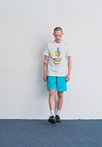 Hvid t-shirt med farverigt grafisk design med en drik; parret med lyse turquoise shorts og sorte sko. Ensfarvet baggrund.