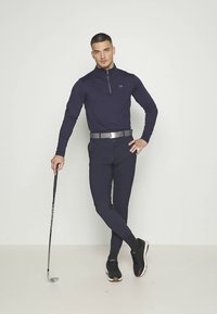 Tamsiai mėlynas, ilgomis rankovėmis su užtrauktuku marškinėliai su CK logotipu, derinami su atitinkančiomis siauro kirpimo kelnėmis ir juodais sportbačiais. Golfo lazdos galas remiasi į jo koją.