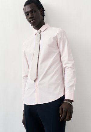 Mężczyzna w jasnoróżowej koszuli i białym krawacie, stojący przy białej ścianie, w czarnych spodniach i srebrnej bransoletce.