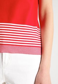 Červený top s texturovaným povrchem, zdobený pruhovaným bílým a červeným lemem. Tkanina je hladká a přiléhavá, spárovaná s bílými kalhotami.