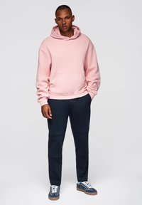 Světle růžová mikina s kapucí a přední kapsou, v kombinaci s tmavě námořnickými zúženými kalhotami a námořnickými teniskami s bílými tkaničkami. Neutrální pozadí.