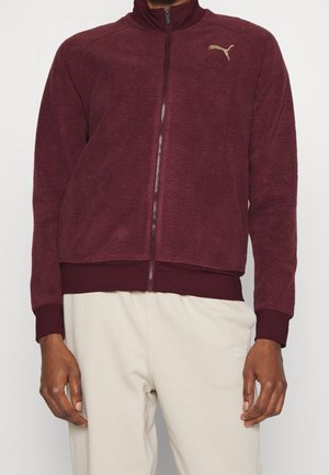 Bordeauxrote Fleecejacke mit durchgehendem Reißverschluss und Logo auf der Brust, getragen zu hellbeiger Hose, gezeigt vom Hals bis zur Hüfte.