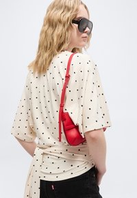 Γυναίκα με κυματιστά ξανθά μαλλιά φορώντας μαύρα γυαλιά ηλίου, κρεμ τοπ με πουά, μαύρο παντελόνι και κόκκινη τσάντα ώμου, γυρισμένη πλάγια με τα χέρια στις τσέπες.