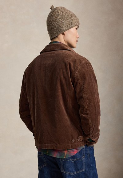 Mand iført en brun manchesterjakke, uldhat med kvast, ternet skjorte og blå jeans, set fra siden mod en ensfarvet baggrund.
