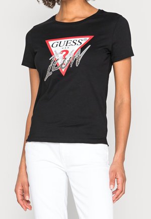 Czarny bawełniany t-shirt z krótkimi rękawami, z czerwonym trójkątnym logo i metalicznym srebrnym napisem. Model nosi białe spodnie.