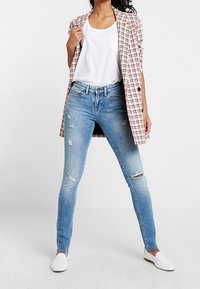 Světle modré potrhané skinny džíny v kombinaci s bílým tílkem a bílou kostkovanou bundičkou. Oblečeno s bílými nazouvacími botami.