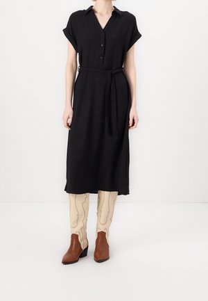 Kobieta w czarnej sukience midi zapinanej na guziki z podwiniętymi rękawami i paskiem, zestawiona z beżowymi, wzorzystymi, sięgającymi kolan kozakami oraz brązowymi botkami na obcasie.