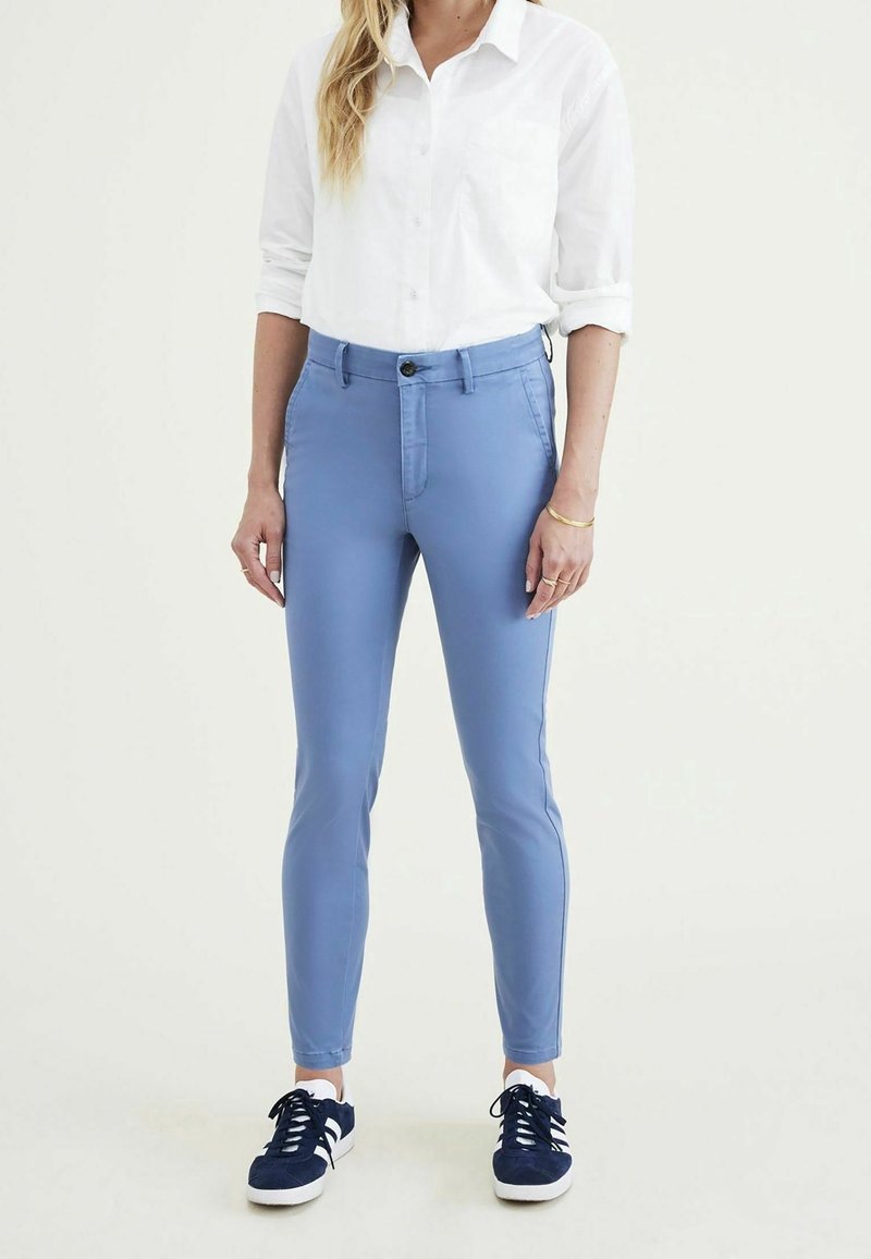 Femme portant une chemise blanche retroussée, un pantalon slim bleu clair et des baskets bleu marine, debout devant un fond uni.