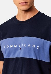 Camiseta de algodón de color azul marino con una franja horizontal de color azul claro con el texto "TOMMYJEANS" en blanco y un pequeño logo. Diseño de mangas cortas y cuello redondo.