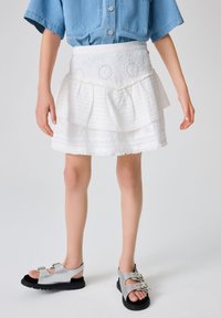 Hvid lagdelt nederdel med tekstureret stof og broderede accenter, kombineret med sølvfarvede sandaler med spænde og en tyk sort sål.