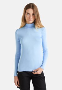 Kobieta z długimi blond włosami, ubrana w jasnoniebieski sweter z golfem i czarne spodnie, stojąca na tle jednolitego, jasnego tła.