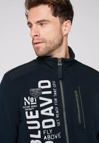 Giacca navy con zip e colletto alto, caratterizzata da stampe in bianco e una tasca con zip. Realizzata in tessuto morbido, con dettagli a vita su spalle.