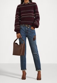 Osoba w bordowym swetrze w wzory, z podwiniętymi niebieskimi dżinsami z łatami, brązowym paskiem, brązowych szpilkach oraz trzymająca brązową skórzaną torebkę.