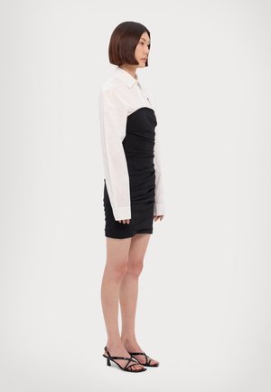 Rochie mini neagră cu volane, cu mâneci lungi albe; are un corset ajustat și o siluetă relaxată, completată de tocuri negre cu barete.