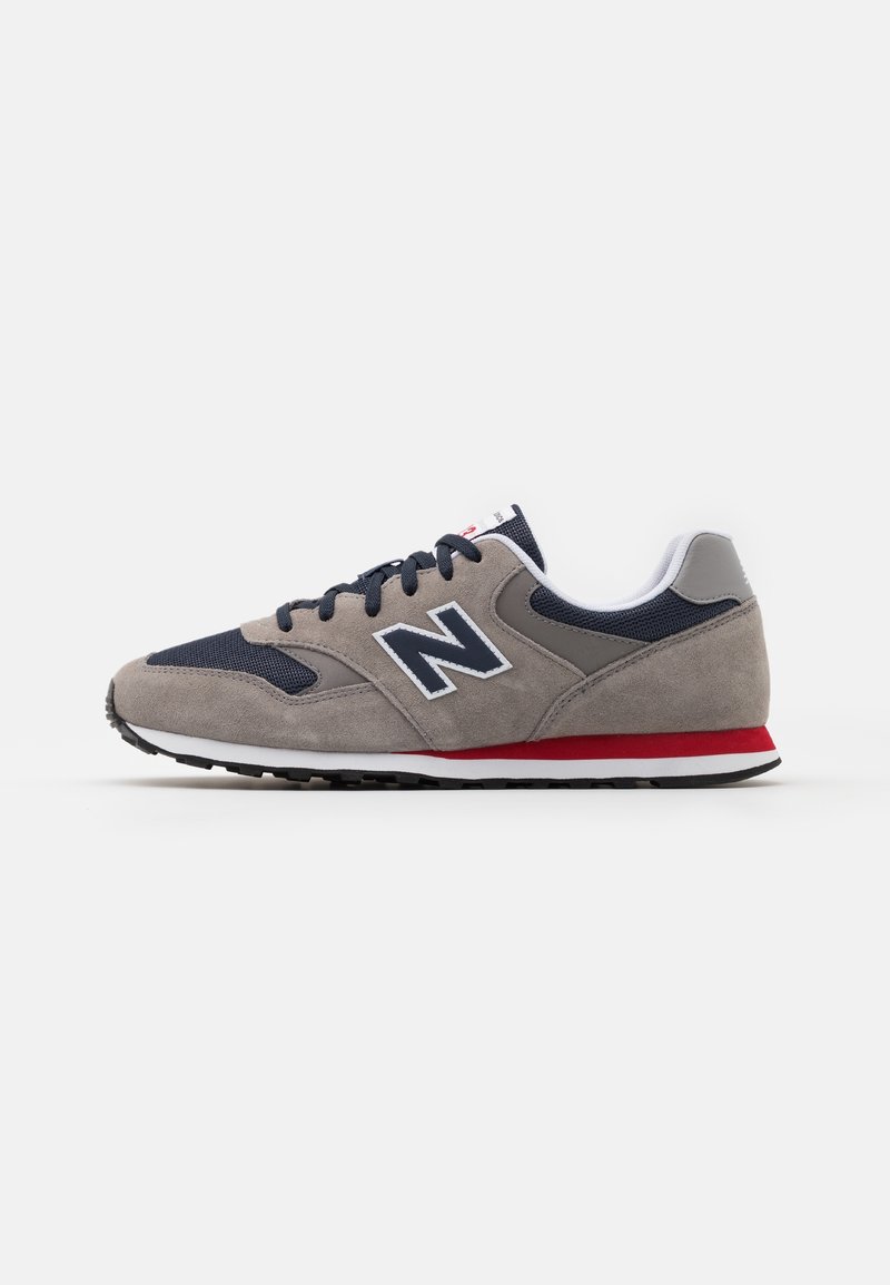 Baskets New Balance gris et bleu marine avec lacets noirs, bord de semelle blanc et bande rouge accentuée, vues de côté sur fond blanc.