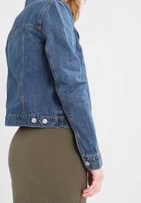 Veste en jean de couleur bleu moyen, avec un design classique, des accents de boutons et une silhouette ajustée, associée à une jupe côtelée olive.
