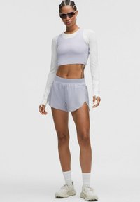 Atletische vrouw in een lichtpaarse crop tanktop, witte langemouwen shirt, bijpassende shorts, witte sokken, sneakers en een zonnebril, staand op een witte achtergrond.
