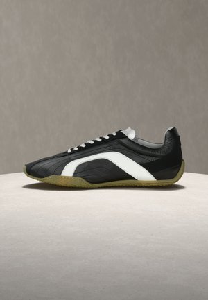 Baskets en cuir noir et blanc avec lacets blancs et semelle texturée vert olive, présentées sur une surface en pierre claire.