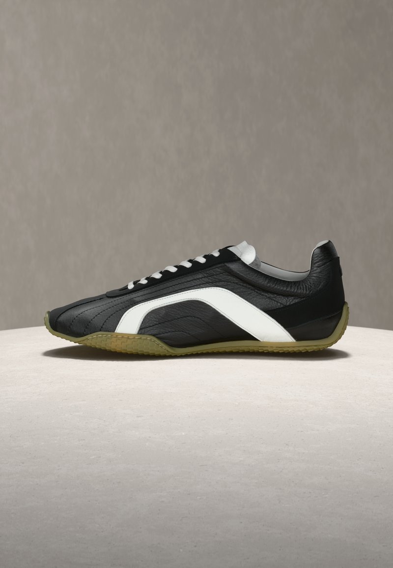 Αθλητικό παπούτσι από ασπρόμαυρο δέρμα με λευκά κορδόνια και σόλα σε ελιά πράσινο με υφή, τοποθετημένο πάνω σε ανοιχτό πέτρινο υπόβαθρο.