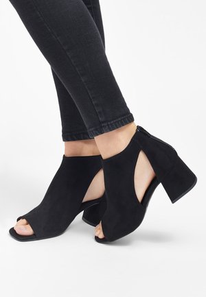 Bottes à talons bloc en suède noir ouvertes à l'avant, avec découpes à la cheville, portées avec un pantalon noir ajusté sur un fond blanc.