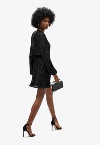 Robe noire à sequins avec des manches longues, taille cintrée et ourlet court. Associée à des talons aiguilles noirs et une pochette noire texturée.