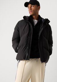 Zwarte pufferjack met gestructureerde accenten, ritssluiting aan de voorkant en capuchon, gecombineerd met een zwart sweater en beige broek met zijstrepen.