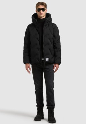 Un mannequin masculin se tient devant un fond gris uni, vêtu d’une doudoune noire à capuche, d’un pantalon noir, de bottes et de lunettes de soleil.