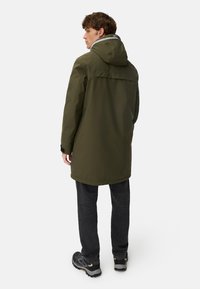 Olivengrüne wasserdichte Parka mit Kapuze, einem Reißverschluss vorne und schwarzen Akzenten. Getragen über dunklen Jeans und grauen Sneakers. Glatte Textur.
