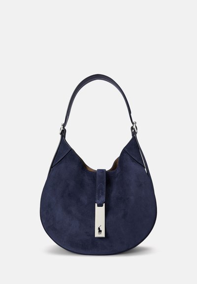 Mörkblå mockaväska med rundad form, silvriga detaljer och övre handtag. Har en vertikal mittaccent och logodetaljer.