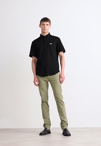 Camicia nera a maniche corte con bottoni e taschino frontale, abbinata a pantaloni slim-fit verde oliva e scarpe nere senza lacci.