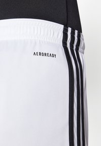 Shorts de sport blancs avec des rayures noires et la marque "AEROREADY" sur le côté gauche. Fabriqués en tissu léger et lisse.