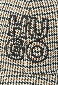 Μαύρη κεντητή λέξη "HUGO" σε μπεζ, μαύρο και λευκό ύφασμα με σχέδιο σκυλοδόντι, πιθανώς μέρος καπέλου ή ενδύματος.