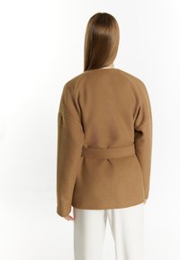 Jachetă din lână de culoare brună, cu un cordon, formă supradimensionată, umeri căzuți și textură netedă. Vedere din spate care arată linii curate și detalii minimale.