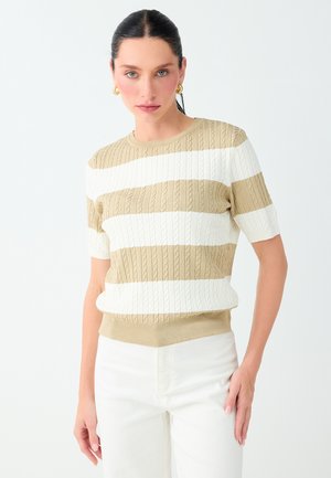 Donna con capelli scuri con maglione a righe beige e bianco a maniche corte in maglia e pantaloni bianchi su sfondo chiaro uniforme.