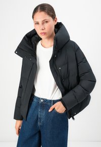 Jeune femme portant une doudoune noire sur une chemise blanche et un jean bleu, debout avec une main dans la poche sur fond blanc.
