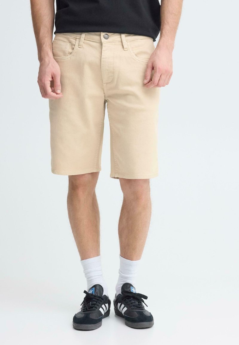 Shorts en denim beige avec une coupe classique, un design à cinq poches, et une fermeture par bouton en métal, assortis à des baskets noires et des chaussettes blanches mi-mollet.