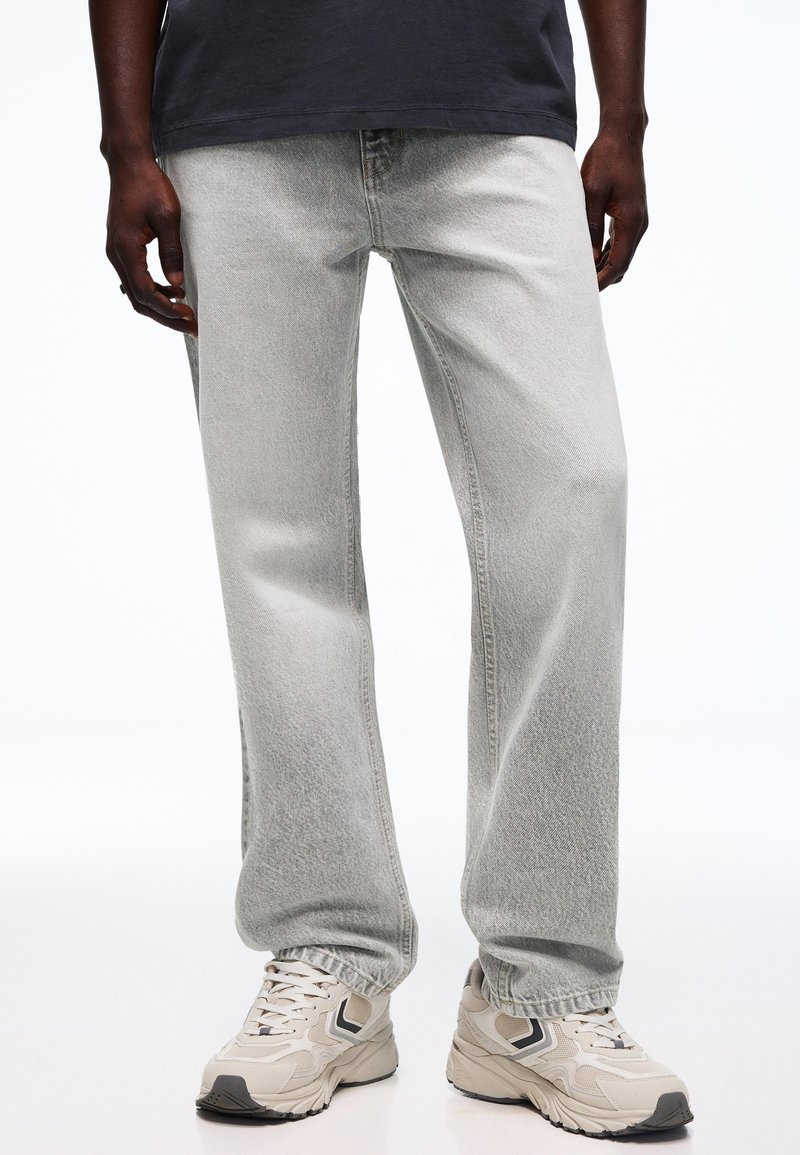 Person trägt hellgraue Jeans mit geradem Bein und beige klobige Sneaker, steht vor einem schlichten weißen Hintergrund.