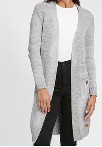Gray knitted cardigan with open front, ribbed cuffs, side pockets, and a branded leather patch, worn over a white top and black pants.