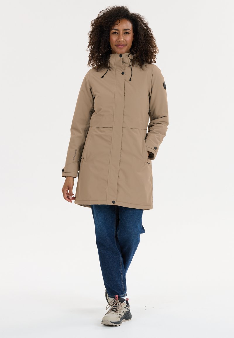 Vrouw met krullend haar, gekleed in een beige knielange jas, blauwe jeans en grijze wandelschoenen, staand tegen een witte achtergrond.
