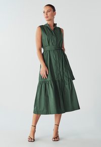 Roheline, varrukateta kleit, millel on krae, vöökoht ja kihiline seelik. Valmistatud siledast kangast, millel on matt viimistlus ja õrn sära.