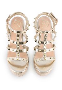 Tacones dorados de punta abierta con múltiples tiras con hebillas que cuentan con tachuelas metálicas. La suela tiene un detalle de yute trenzado, ofreciendo un contraste texturizado.