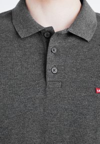 Polo gris en tissu texturé, doté d'un col classique, de trois boutons foncés et d'un petit logo rouge sur la poitrine.