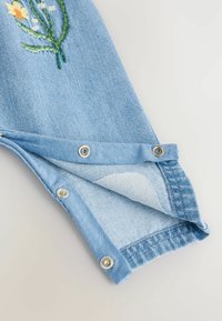 Tissu en denim de couleur bleu clair avec des broderies florales. Comprend des boutons pression au niveau des poignets et une texture douce.