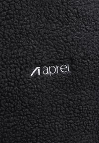 Juodos tekstūros fliso audinys su balta siuvinėta „aprel“ logotipu, esančiu kadro centre.