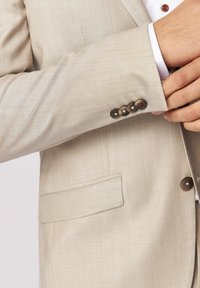Hellbeige Anzugjacke mit glatter Textur, die einen geschwungenen Kragen, eine Brusttasche und vier dunkle Knopfakzente am Ärmel aufweist.