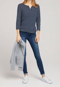Mørkeblå langærmet top med tekstureret mønster og henley-udskæring; parret med mørke skinny jeans og hvide sneakers, mens der holdes en grå blazer.