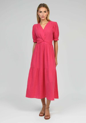 Femme portant une robe midi rose vif avec des manches bouffantes, un col en V et un détail noué à la taille, associée à des escarpins marron ouverts à l'avant.