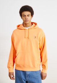 Oranje hoodie met een buidelzak, verstelbare trekkoord capuchon en een klein logo-patch op de borst. Zacht materiaal en geribbelde manchetten.
