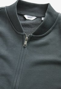 Sivi zip-up dukserica s rebrastim ovratnikom i srebrnim patentnim zatvaračem. Tkanina ima glatku teksturu i jednostavan je dizajn bez uzoraka.